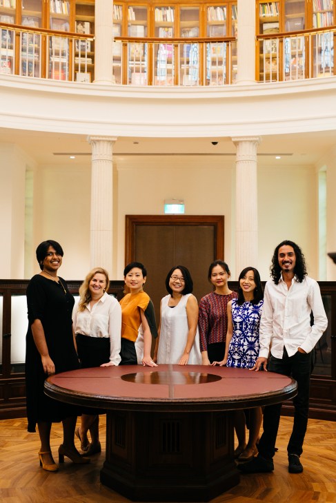 Seven museum curators standing around a large circular wooden table in a neoclassical gallery space with columns and upper-level wooden-framed windows.