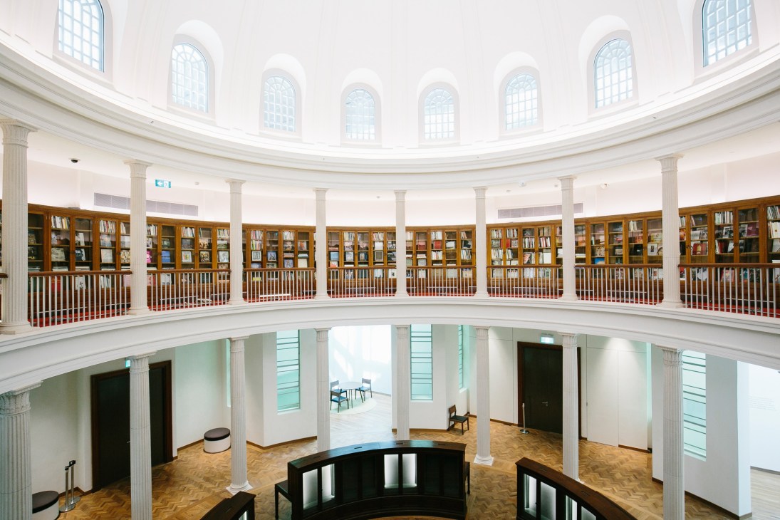 Circular reading room with arched windows, white columns, wooden bookshelves, and natural light from above.