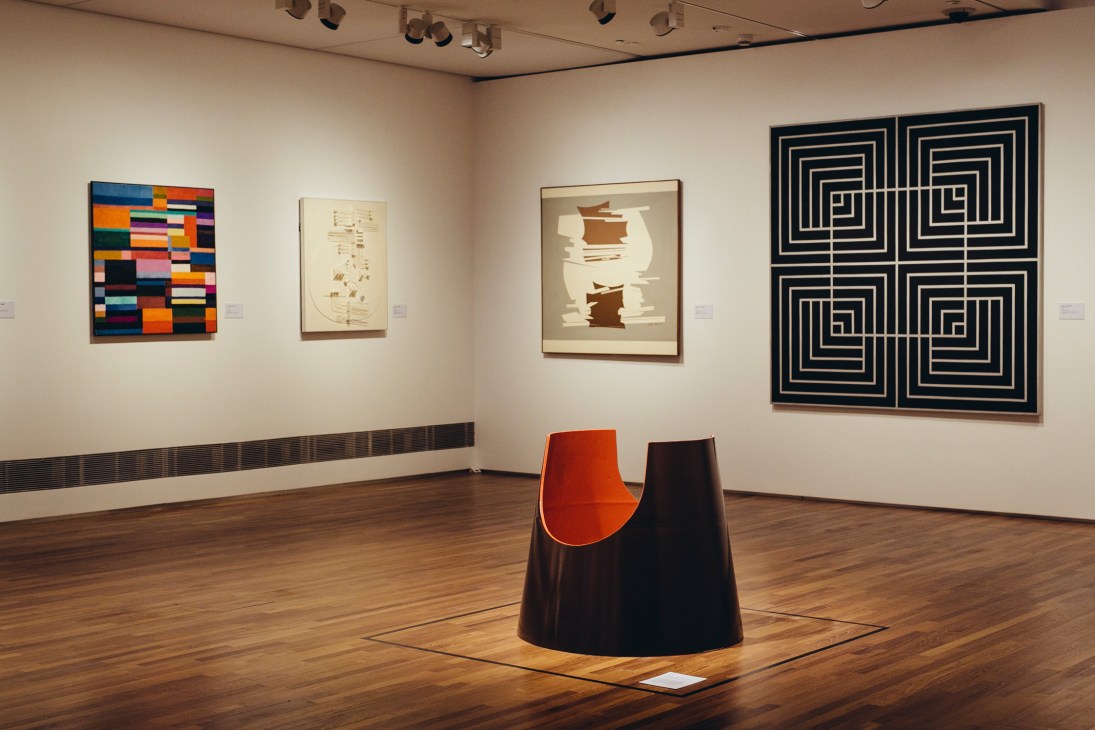 Gallery interior displaying contemporary artworks including colorful abstract paintings and geometric prints on white walls with wooden flooring.
