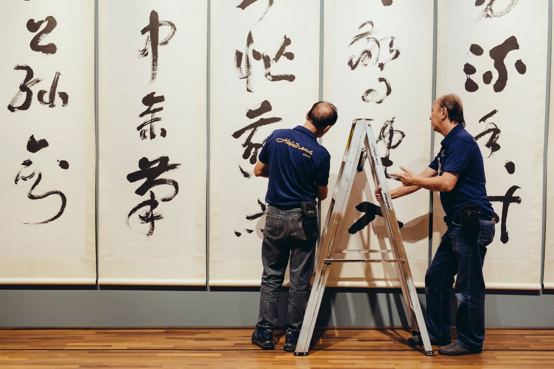 Two technicians on a ladder aligning a large calligraphic painting on a gallery wall.