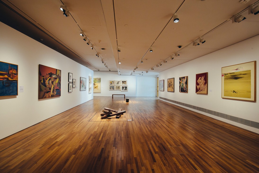 Spacious gallery interior with wooden flooring, white walls displaying various artworks, and sculptural pieces on the floor.