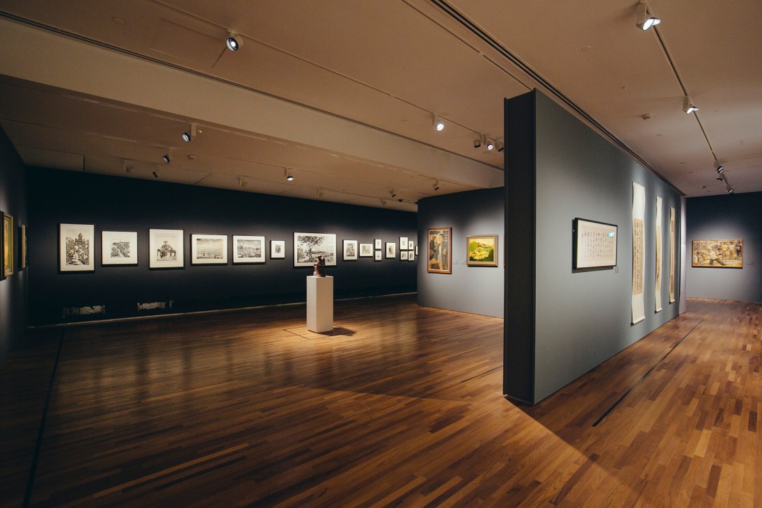 Gallery interior with artworks displayed chronologically on dark and light walls with wooden flooring.