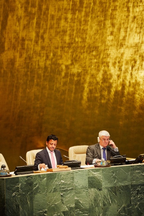 Two delegates sit at a curved desk in the UN General Assembly Hall with microphones and water glasses.
