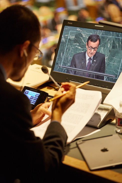 A UN interpreter views a live feed of a delegate speaking while monitoring interpretation equipment and taking notes.
