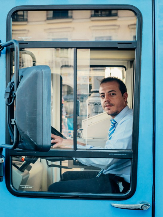Madrid EMT bus driver Luis at the helm of a blue electric microbus, wearing a light blue shirt and striped tie.