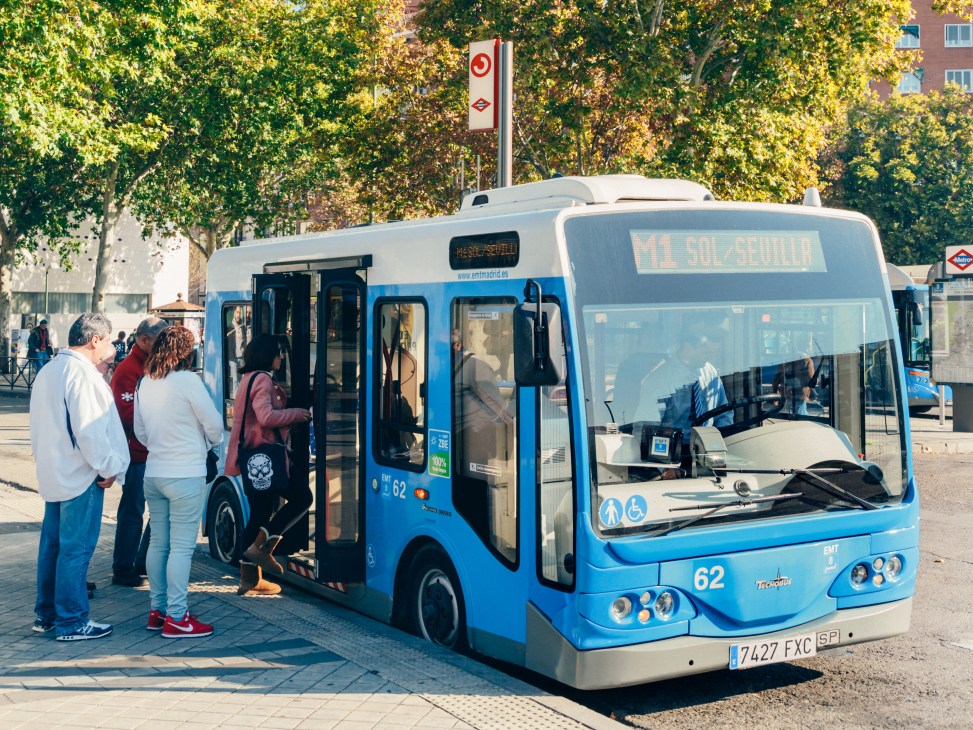 Passengers boarding a blue and white EMT electric microbus on a Madrid street.