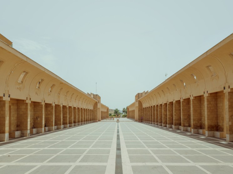 Symmetrical sand-colored stone colonnade with arched openings flanking a geometric paved courtyard under pale sky