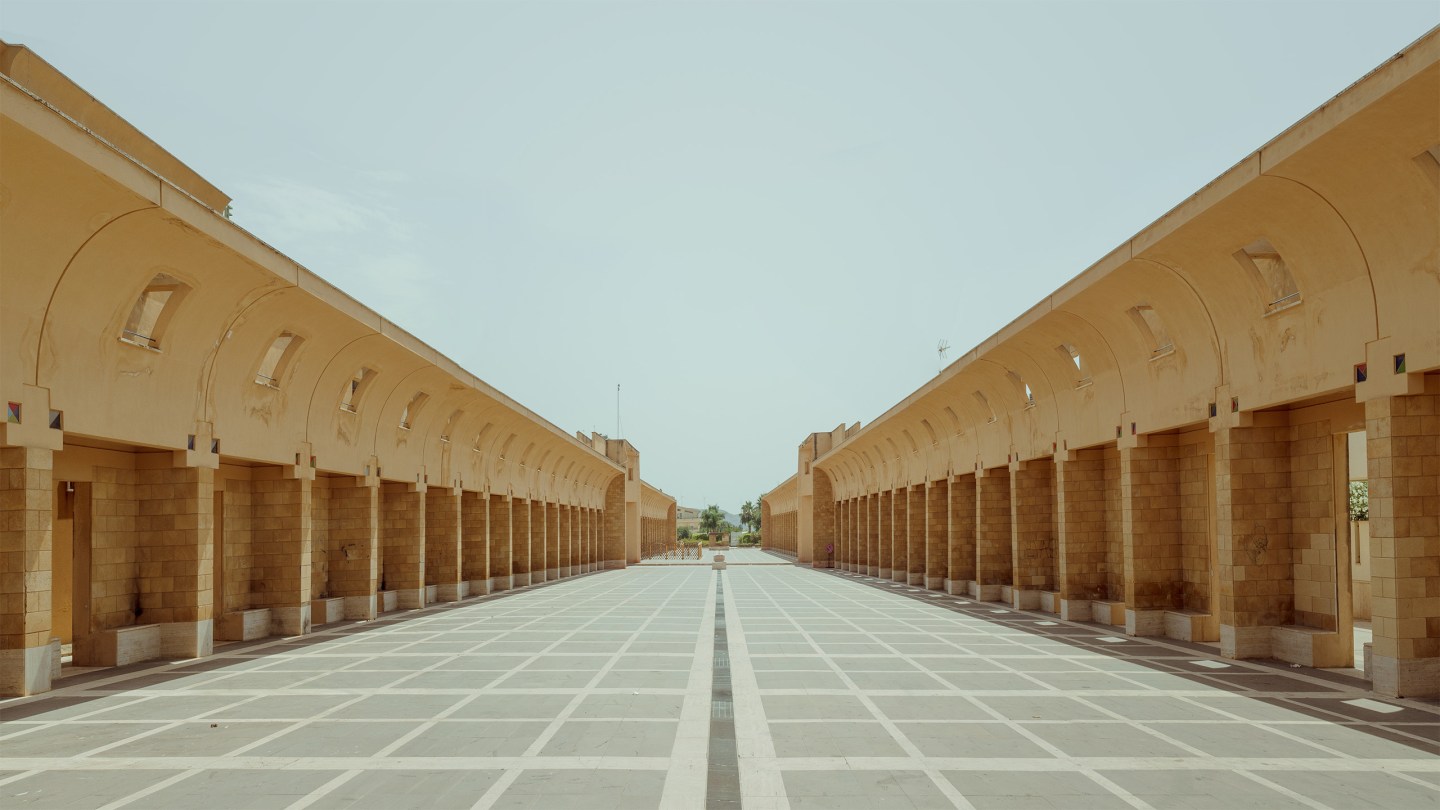 Symmetrical sand-colored stone colonnade with arched openings flanking a geometric paved courtyard under pale sky