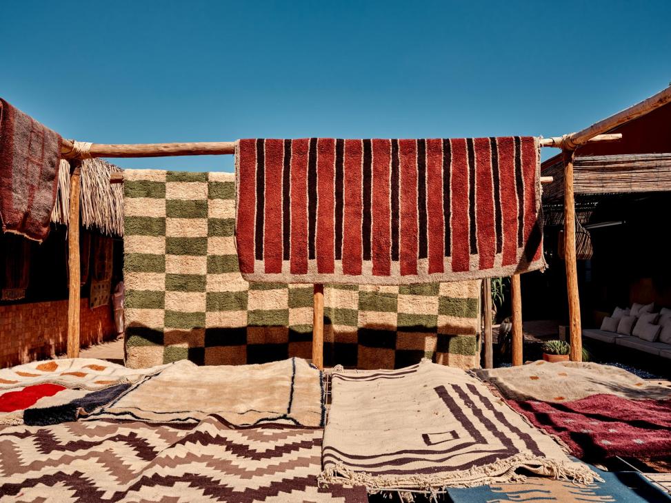 Moroccan rugs in red, green, and cream patterns hang and dry on wooden frames under a bright blue sky at a Beni Rugs facility.