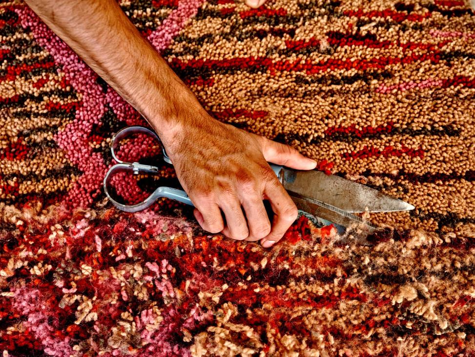 A hand holding scissors trims a colorful Beni Rugs handwoven rug with red, pink, and cream tones.