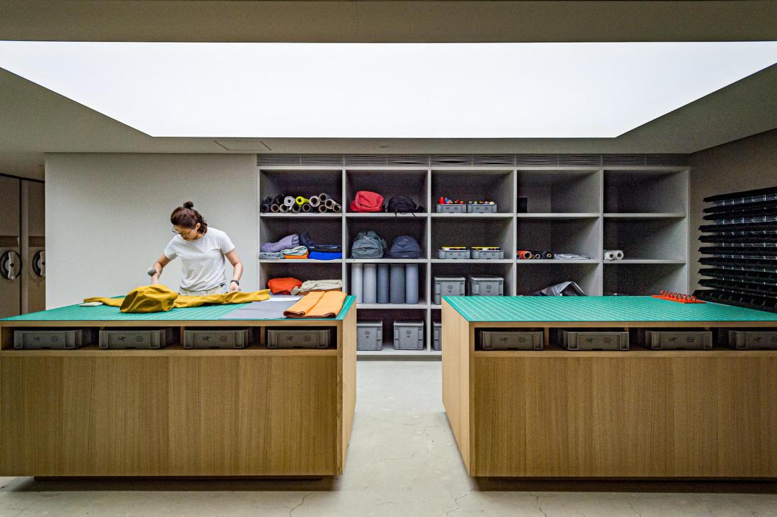 Woman in white t-shirt working with yellow fabric on a turquoise cutting table in a bright, minimalist workshop with shelving and materials.