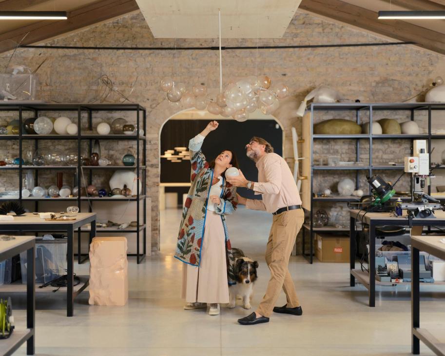 Two people examine a hanging glass light fixture in a workshop with shelving and materials visible.