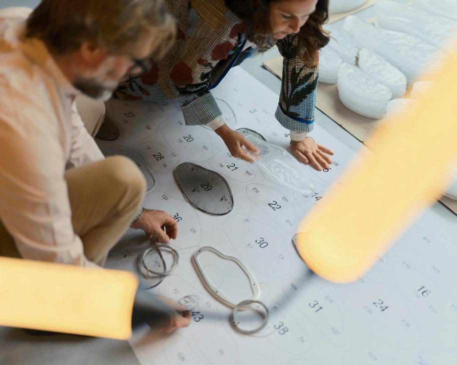 Two people examining light fixture components and design sketches on a table with a calendar layout.