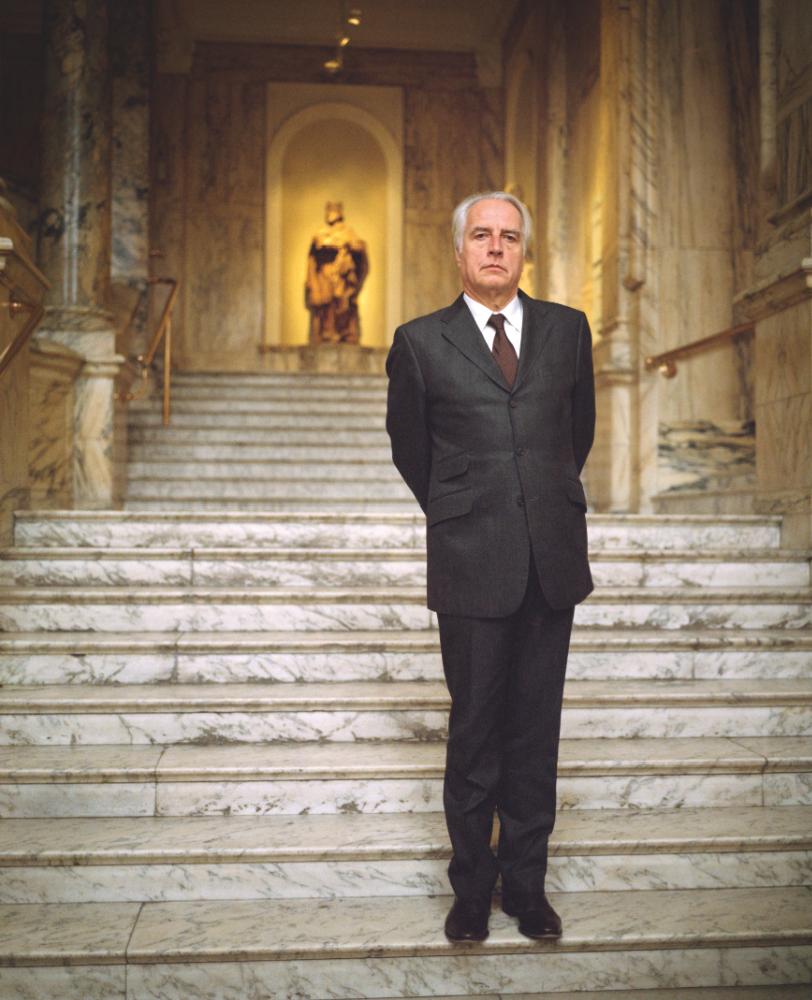 Martin Roth, museum director, stands in a formal black suit on a grand marble staircase in an elegant institutional hallway.