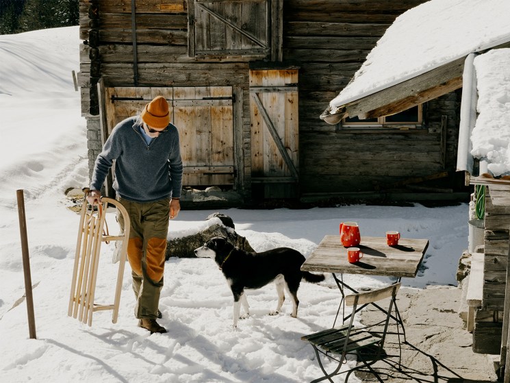 Man walks past snowy cabin holding a sledge