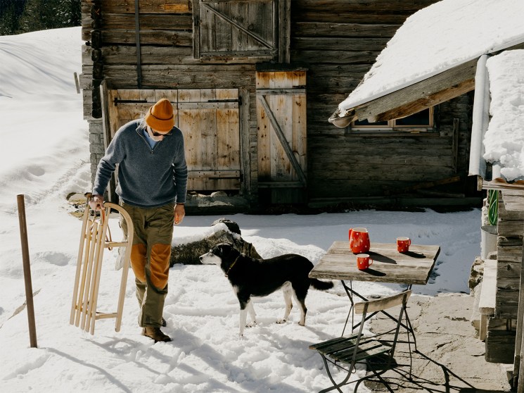 Man walks past snowy cabin holding a sledge