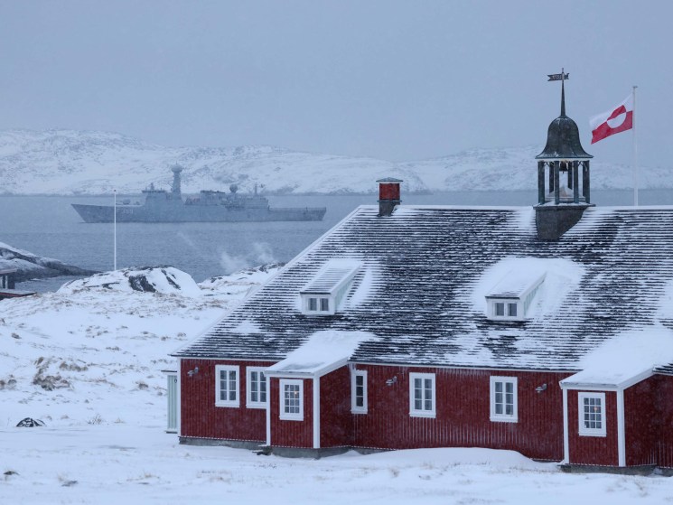 NUUK, GREENLAND - JANUARY 18: The Greenlandic flag flies over a building as the HDMS Vaedderen frigate of the Danish Navy patrols behind on January 18, 2026 in Nuuk, Greenland. Greenlandic, Danish and other European leaders are hoping they can still avert an intervention by the United States to forcefully acquire the island as U.S. President Donald Trump continues to insist the U.S. must have Greenland, suggesting even by military means if necessary. (Photo by Sean Gallup/Getty Images)