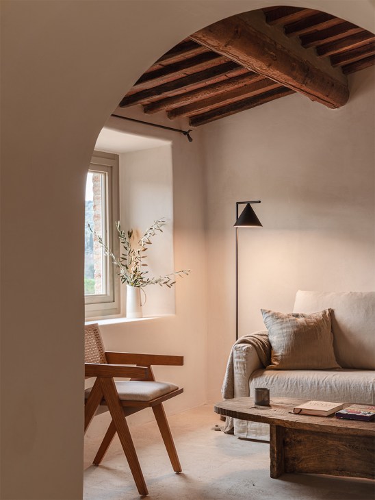 Minimalist hotel room with exposed wooden ceiling beams, white walls, wooden chair, sofa with cushions, black floor lamp, and window with dried flowers.