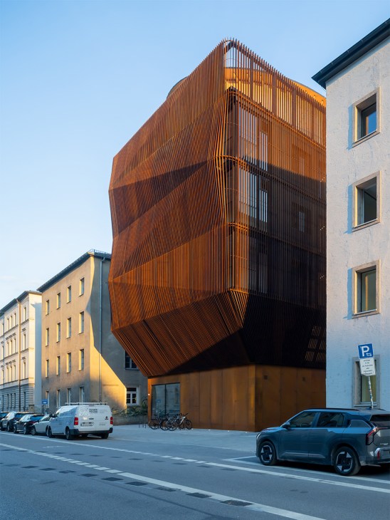 Modern five-story timber building with distinctive corrugated copper-brown facade sitting between traditional residential buildings on an urban street.
