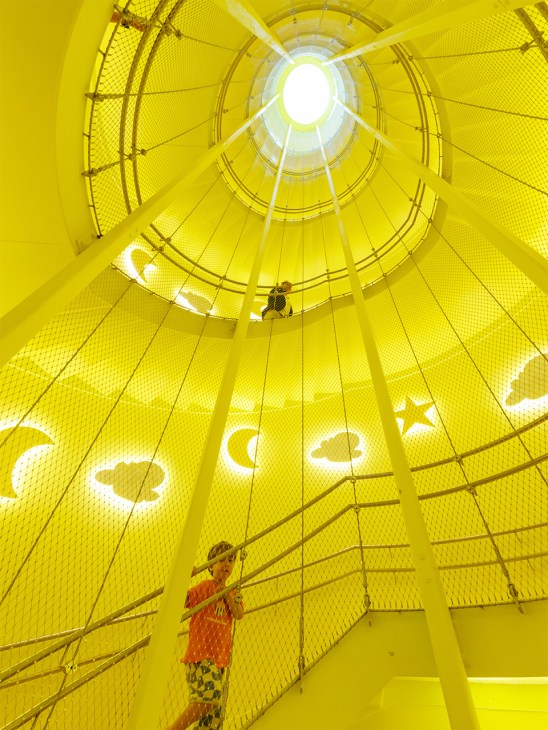 Bright yellow spiral staircase inside the Kinderoase daycare centre with geometric wall patterns and a child climbing near the top.