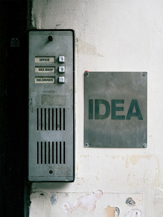 Weathered intercom panel with buttons labeled Office, Idea Shop, and Deliveries next to a metal plaque reading Idea mounted on a white wall.