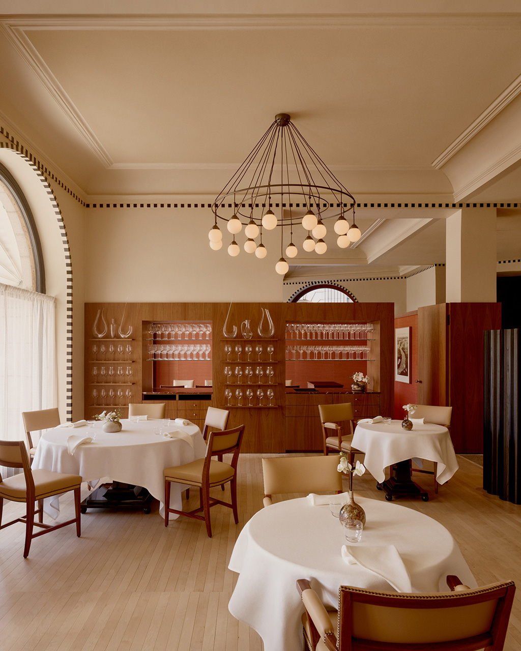 Fine dining restaurant interior with round tables, wooden chairs, pendant chandelier, and glass display cabinetry.