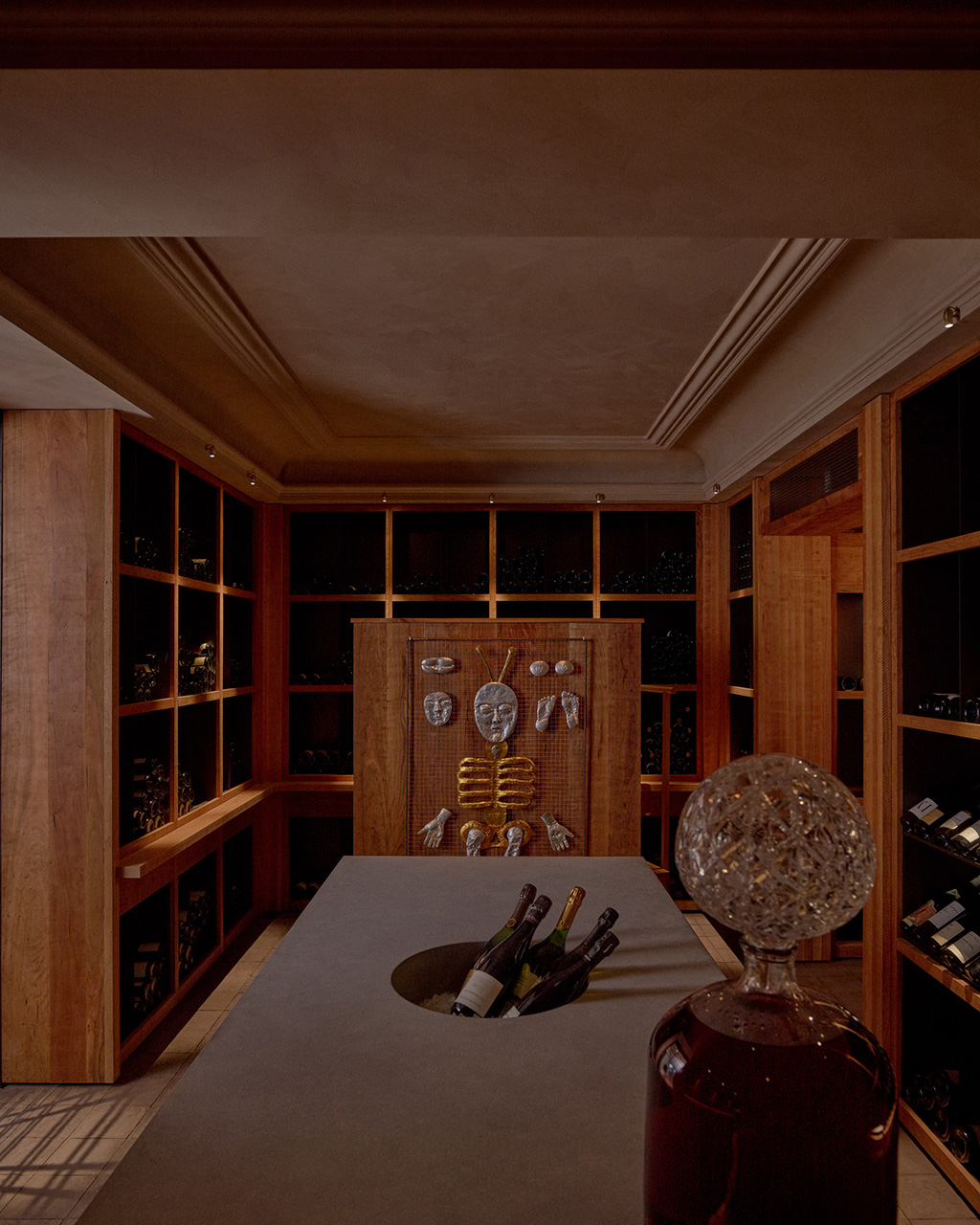 A luxurious wine cellar with wooden cabinetry, glass displays, a central tasting table with wine bottles, and decorative sculptural elements.
