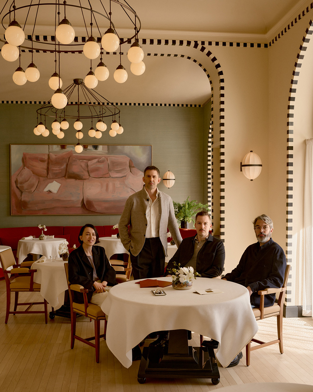 Four people stand and sit around a white-draped circular table in a modern restaurant with geometric pendant lights and striped archways.