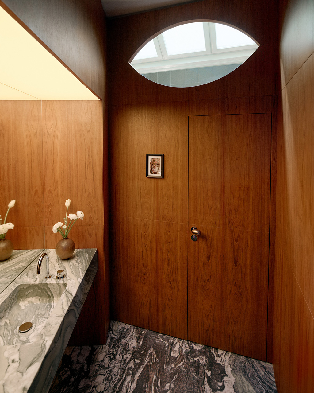 Modern bathroom with warm wood paneling, marble sink countertop, oval skylight, and white flowers in a vase.