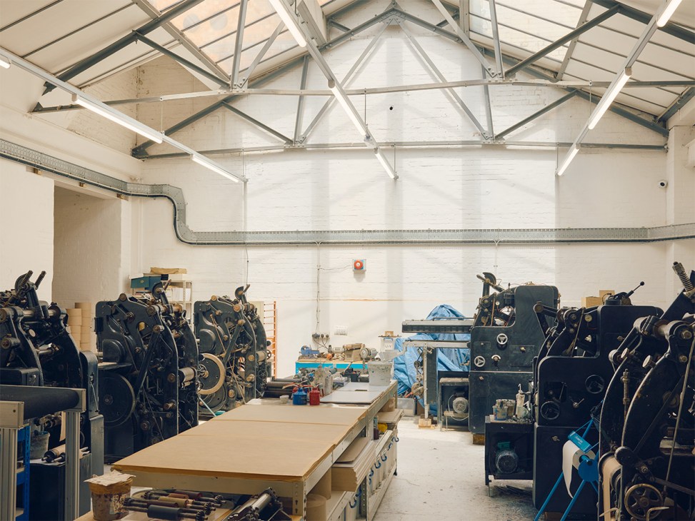 Industrial printing machinery and equipment arranged in a bright warehouse space with metal roof trusses and skylights.