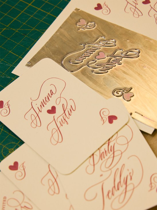 Handwritten wedding invitations with gold foil lettering and decorative hearts on a cutting mat.