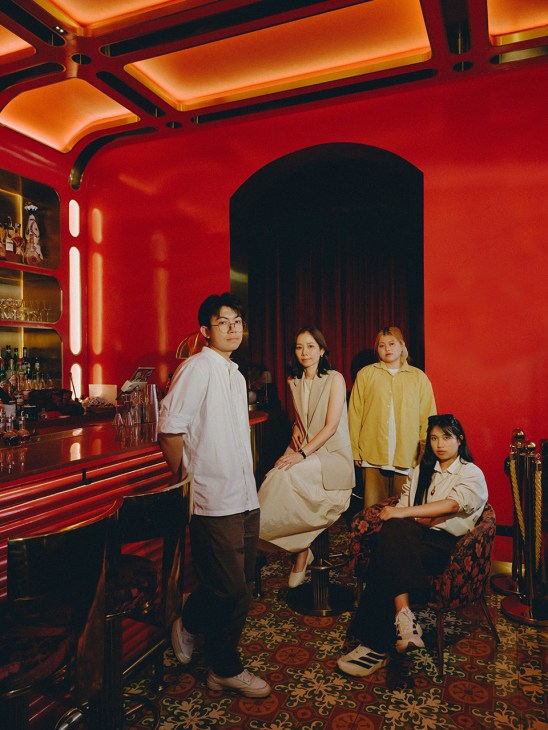 Four people pose in a red-walled bar with neon lighting and ornate patterned flooring.