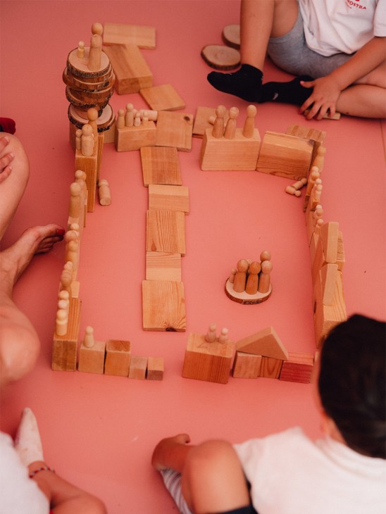 Wooden building blocks and peg figures arranged on a pink surface with children's hands visible around the edges.