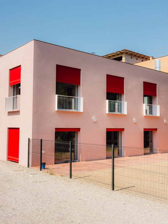 Exterior of Escola Casa Nostra kindergarten with dusty pink facade, red window awnings, white metal railings and black security fencing.