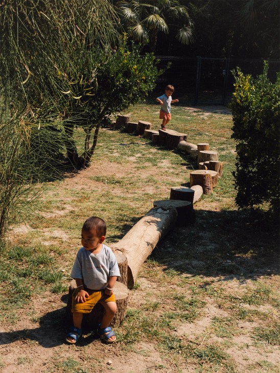 Young children playing and balancing on a line of wooden logs in a garden at Escola Casa Nostra kindergarten.