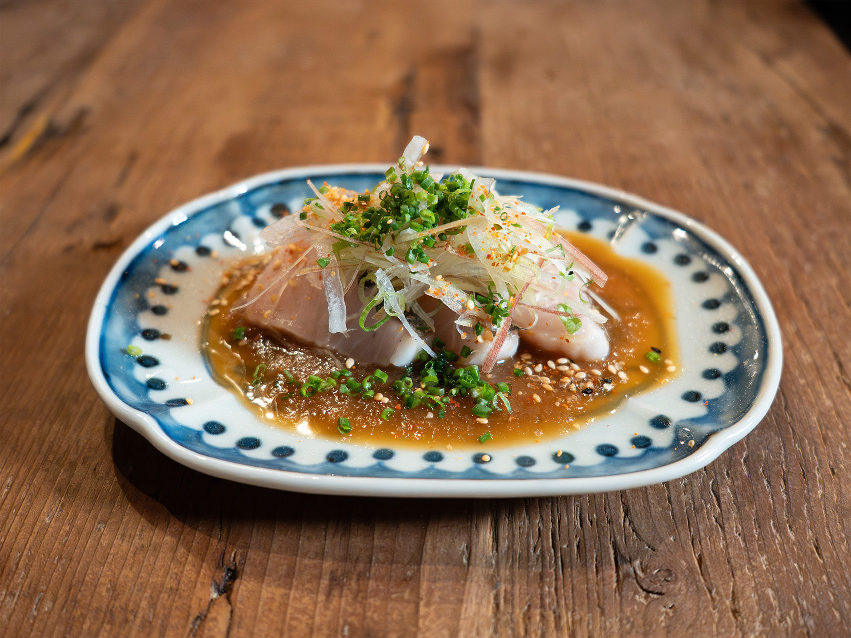 A delicate fish fillet topped with julienned vegetables and microgreens on a blue and white patterned plate with amber-colored sauce.