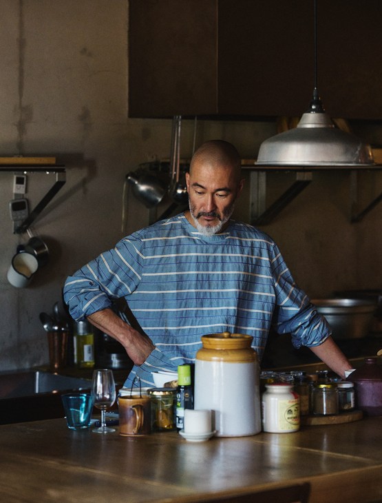 Chef Shinichiro Harakawa wearing a blue striped Prada overshirt in a kitchen with pendant lights and jars.