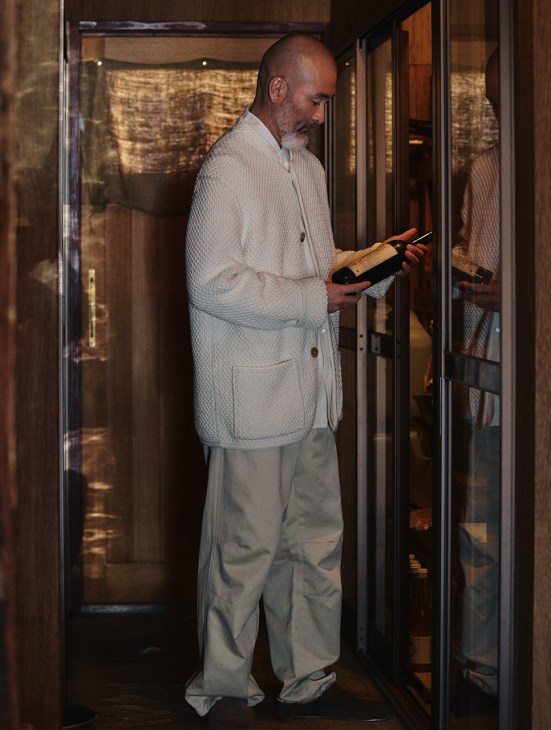 Man in cream quilted Loro Piana jacket and trousers holding wine bottle in doorway.