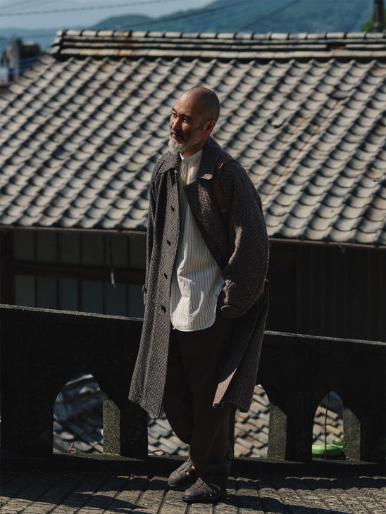 Chef Shinichiro Harakawa wearing a Yoke coat and Visvim shirt stands on a traditional tiled rooftop overlooking mountains.