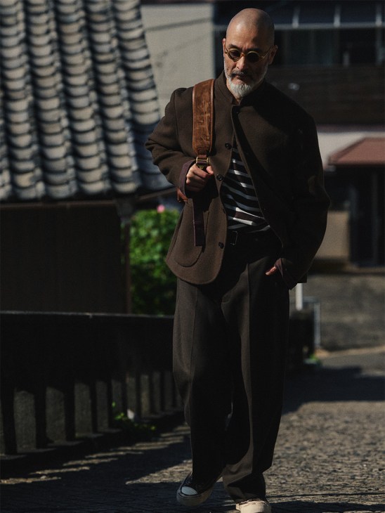 Man wearing Officine Générale jacket and trousers with Outil long-sleeved T-shirt, Visvim trainers, Porter Classic glasses and Bennett Winch backpack on a city street.