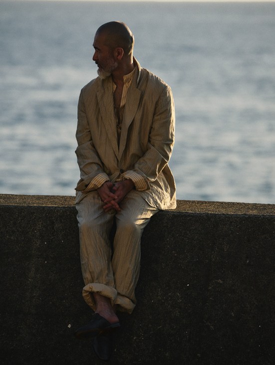 Chef Shinichiro Harakawa sits on a stone wall overlooking the sea, wearing a Zegna jacket, shirt, trousers and shoes.
