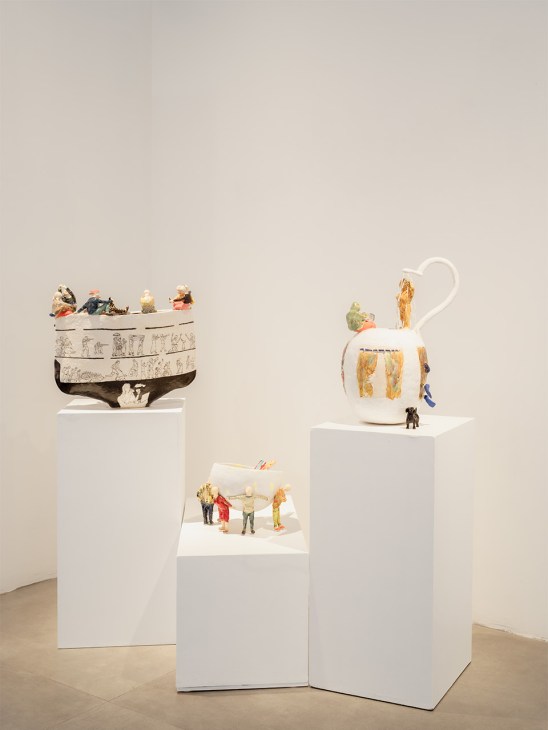 Three white ceramic vessels with gold accents displayed on gallery pedestals, featuring miniature figures.