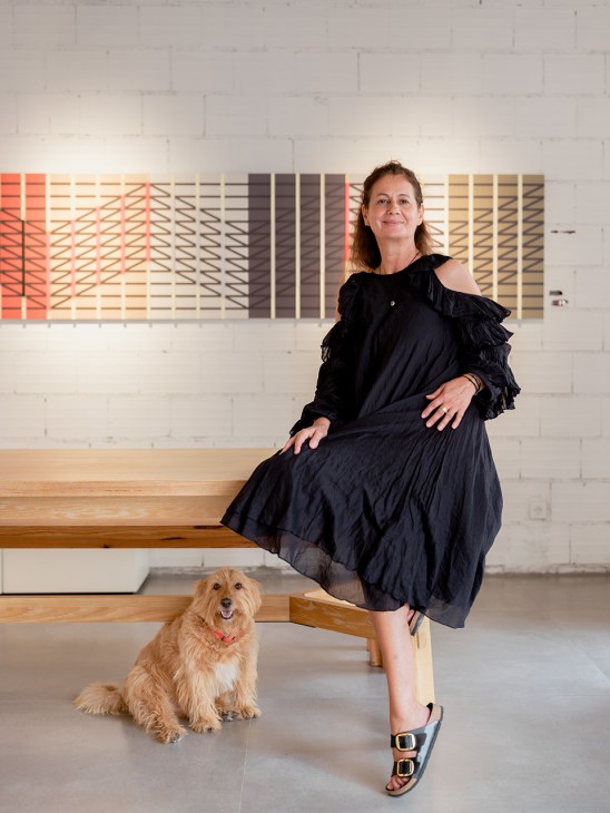 Maria Assunção Albuquerque, curator of Módulo Twist, sits on a wooden bench with her dog Hashi in front of a white brick wall with a colorful geometric light installation.