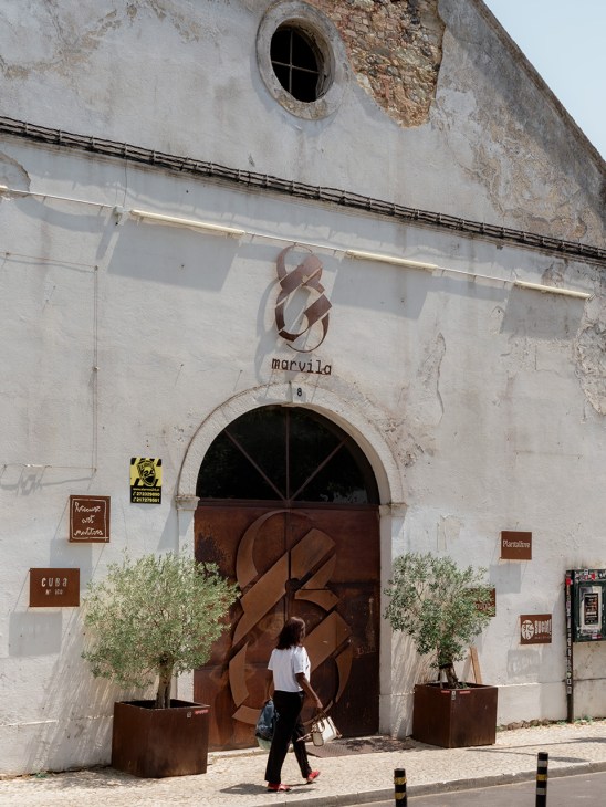 Weathered white building façade with brown wooden arched door, olive plants, and Marvila signage above.