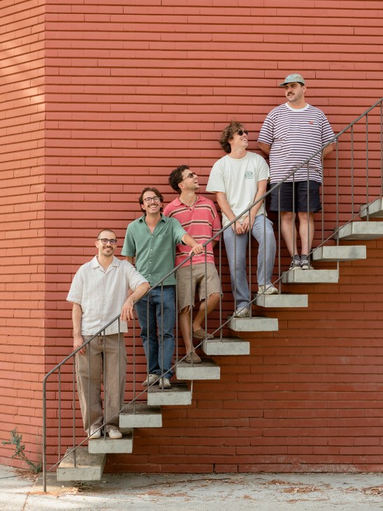 Five members of Portuguese band Napa pose on concrete steps against a red brick wall.