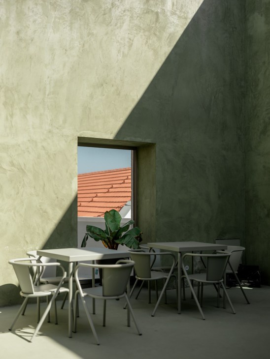 Minimalist indoor courtyard with white chairs at tables, concrete walls, and a window framing orange roof tiles and green plant.