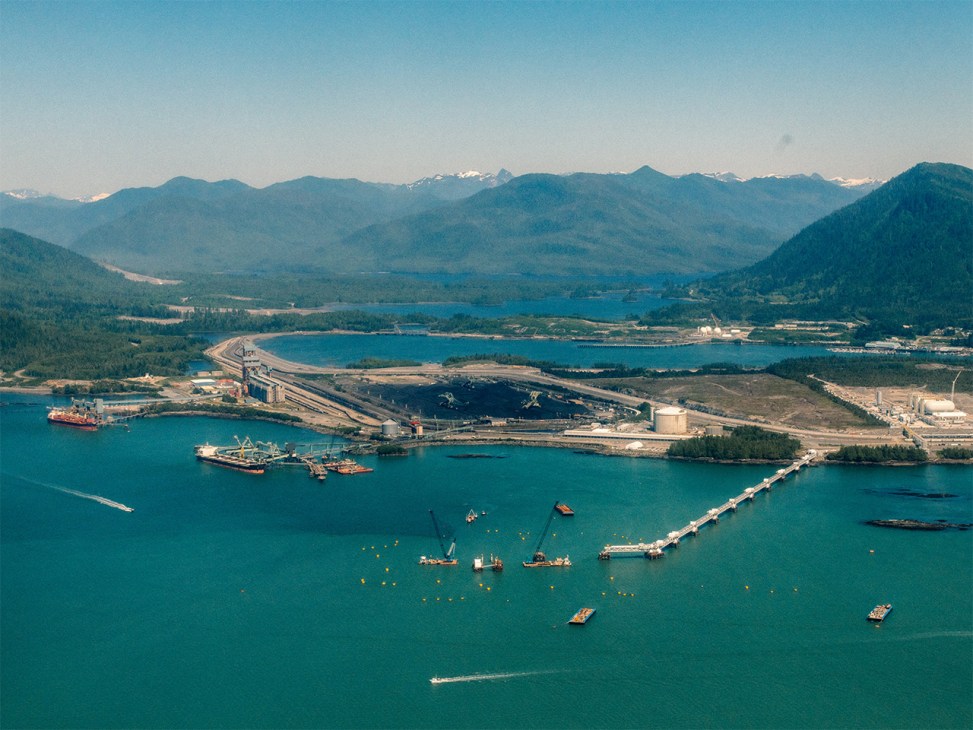 Aerial view of Ridley Island port facility with container ships, industrial infrastructure, and surrounding forested mountains and lakes.