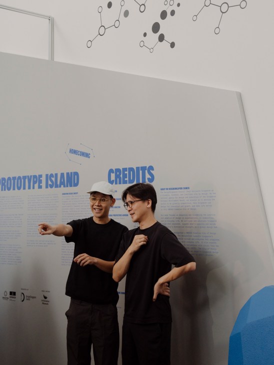Two men in black shirts and glasses stand in front of a wall with blue text reading 'PROTOTYPE ISLAND' and 'CREDITS', with molecular diagrams on the ceiling above.