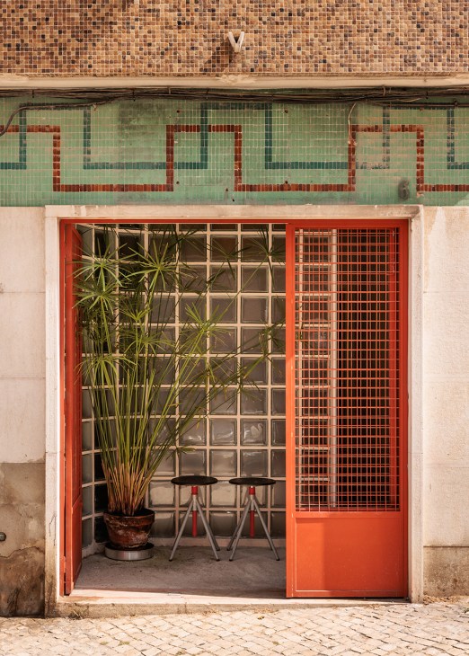 Red-framed shopfront with orange metal grille door and glass blocks, green tiled above, potted plant and chairs visible inside.
