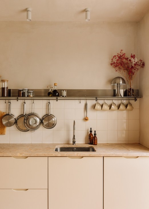 Minimalist kitchen with beige cabinetry, wooden countertop, metal shelving with hanging pans and cups, white subway tile backsplash, and dried flowers in a vase.
